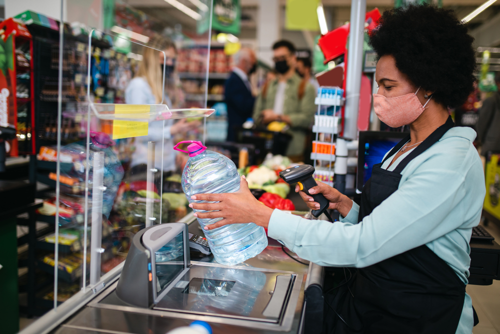 Woman with a face mask on scanning items at a retail store behind a plexiglass window.