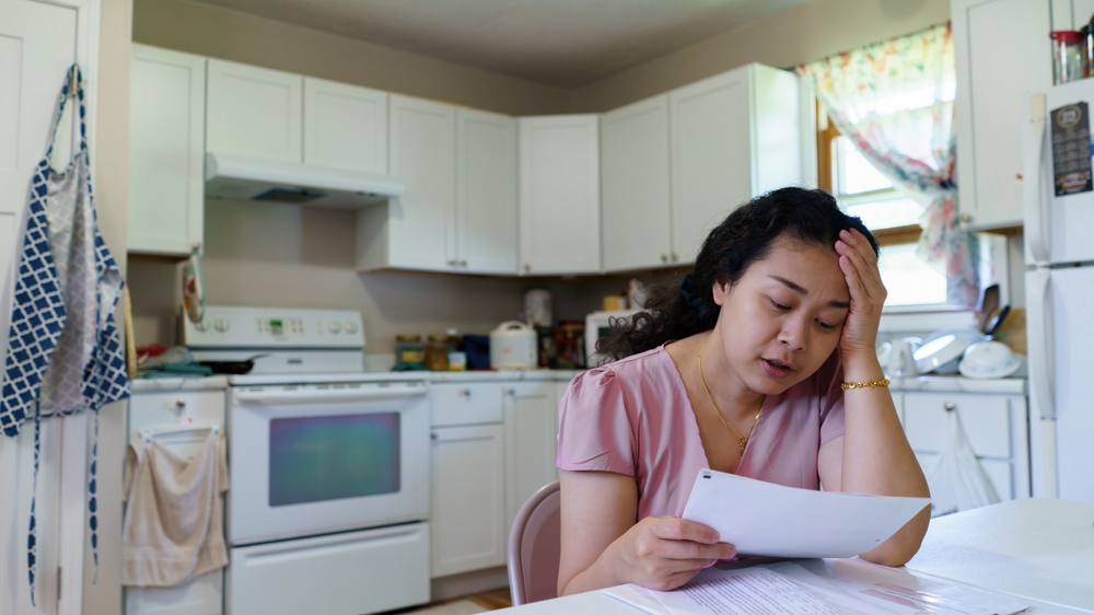 A woman of Asian descent sits in a kitchen looking distressed, with her head in her left hand and a bill in the other. 